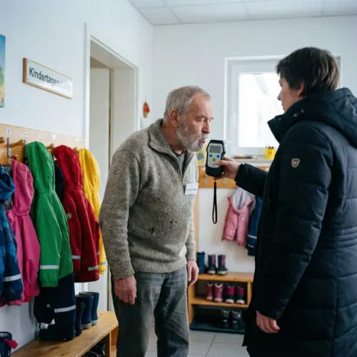 Vor einer bunten Kita-Garderobe bläst eine ältere Frau ernst in ein ihr entgegengehaltenes technisches Messgerät.